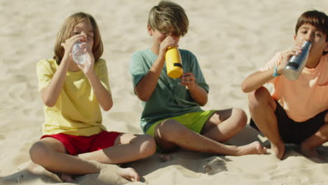 Niños-Sentados-En-La-Playa-Y-Bebiendo-Agua-Después-De-Jugar-Al-Fútbol.